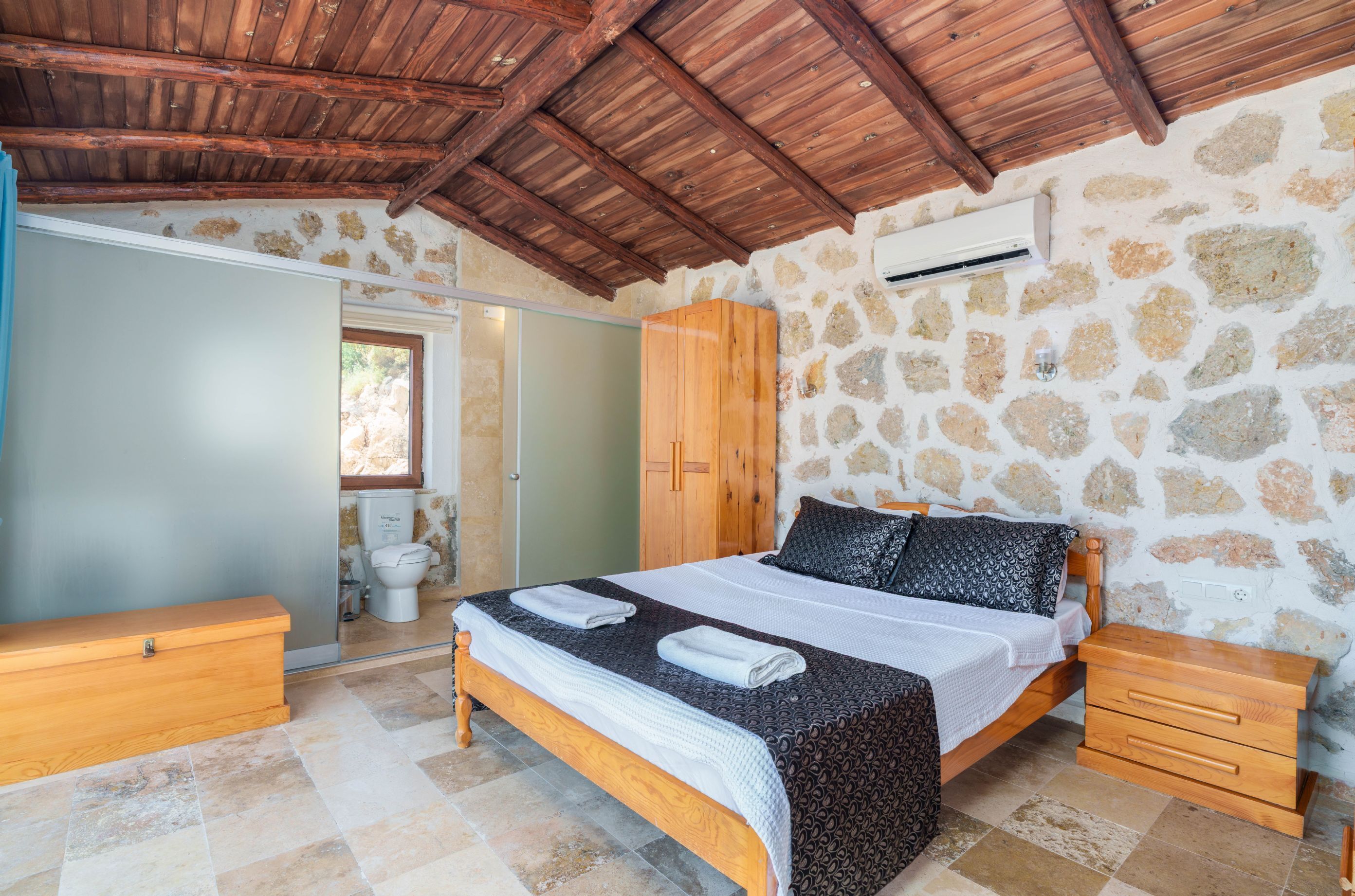 A bedroom with a stone wall, wooden ceiling, a bed with a patterned bedspread, wooden nightstands, and a wooden wardrobe.