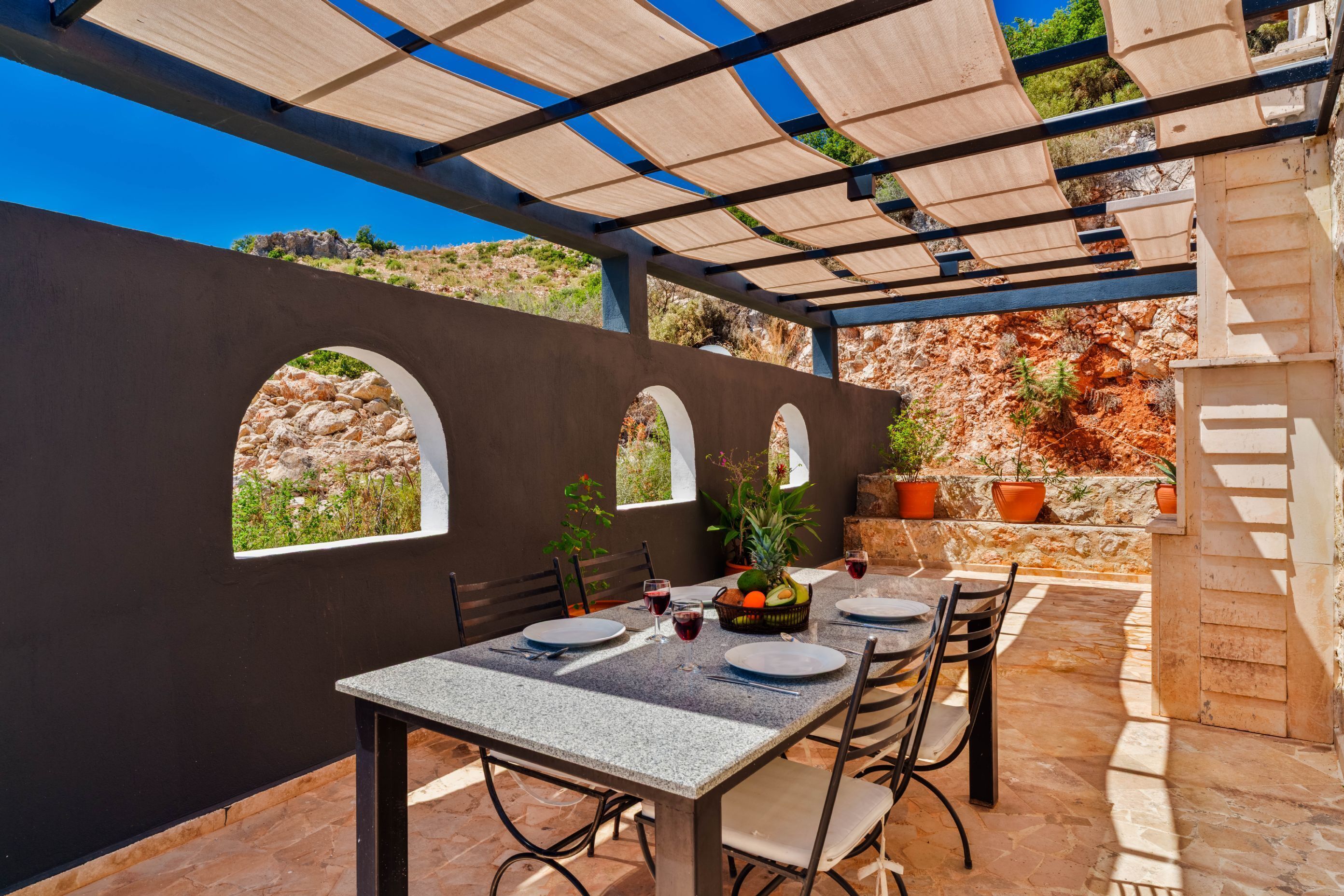 An outdoor dining area with a table set for four, surrounded by chairs, under a shaded pergola.