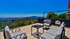 Eine Terrasse mit Tisch und Stühlen, Blick auf das Meer und eine gebirgige Landschaft.