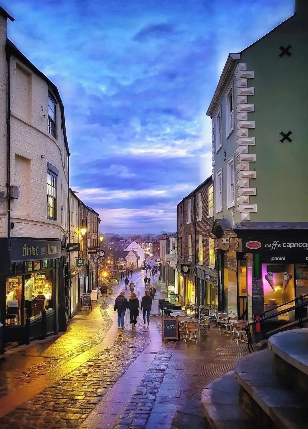 A cobblestone street lined with shops and cafes, with people walking and a sunset in the background.
