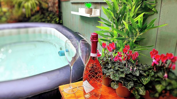 A hot tub with a bottle of rose Prosecco and two glasses on a wooden table, surrounded by potted plants and flowers.