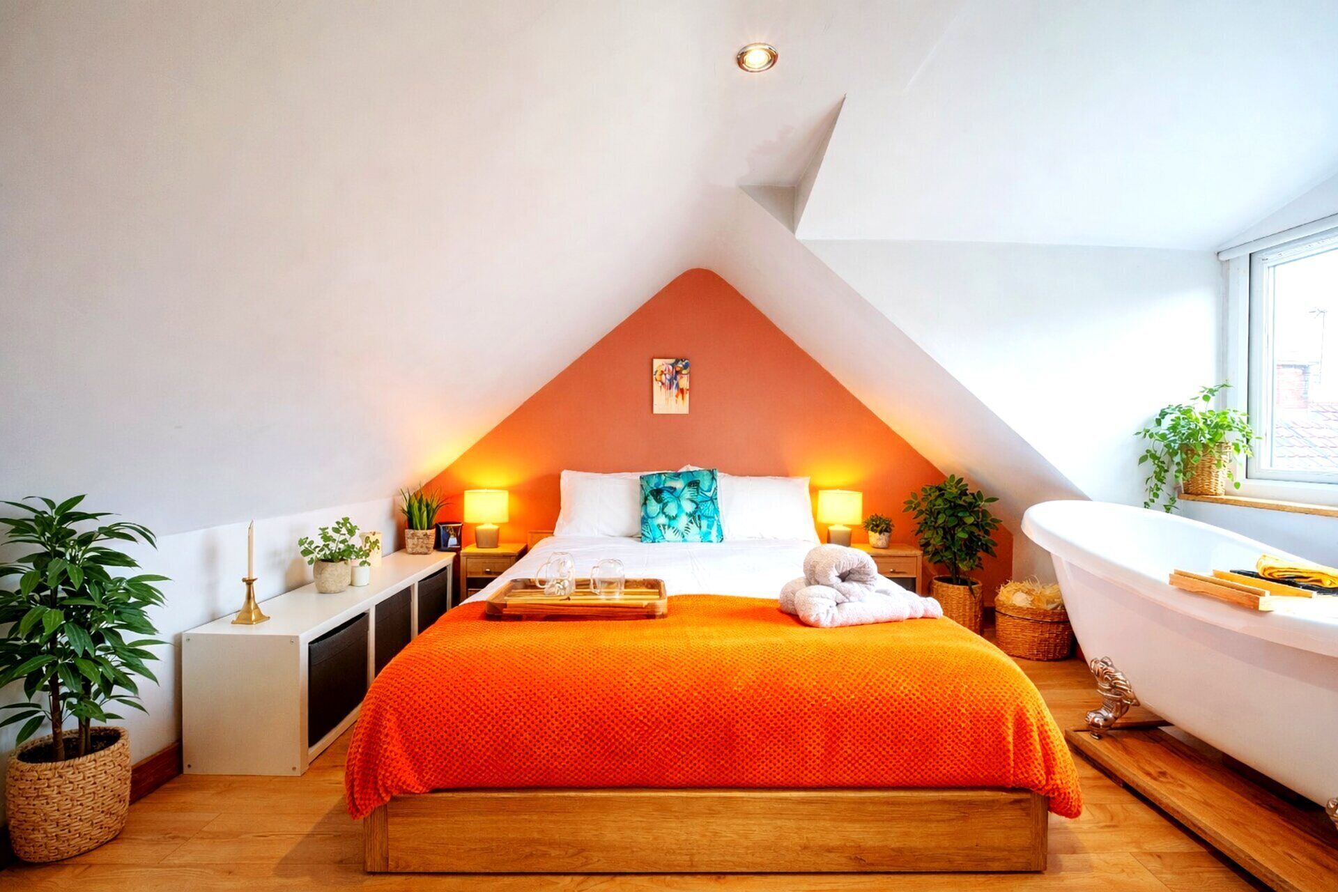A bedroom with a sloped ceiling, a large bed with an orange bedspread, bedside lamps, a white bathtub, and potted plants.