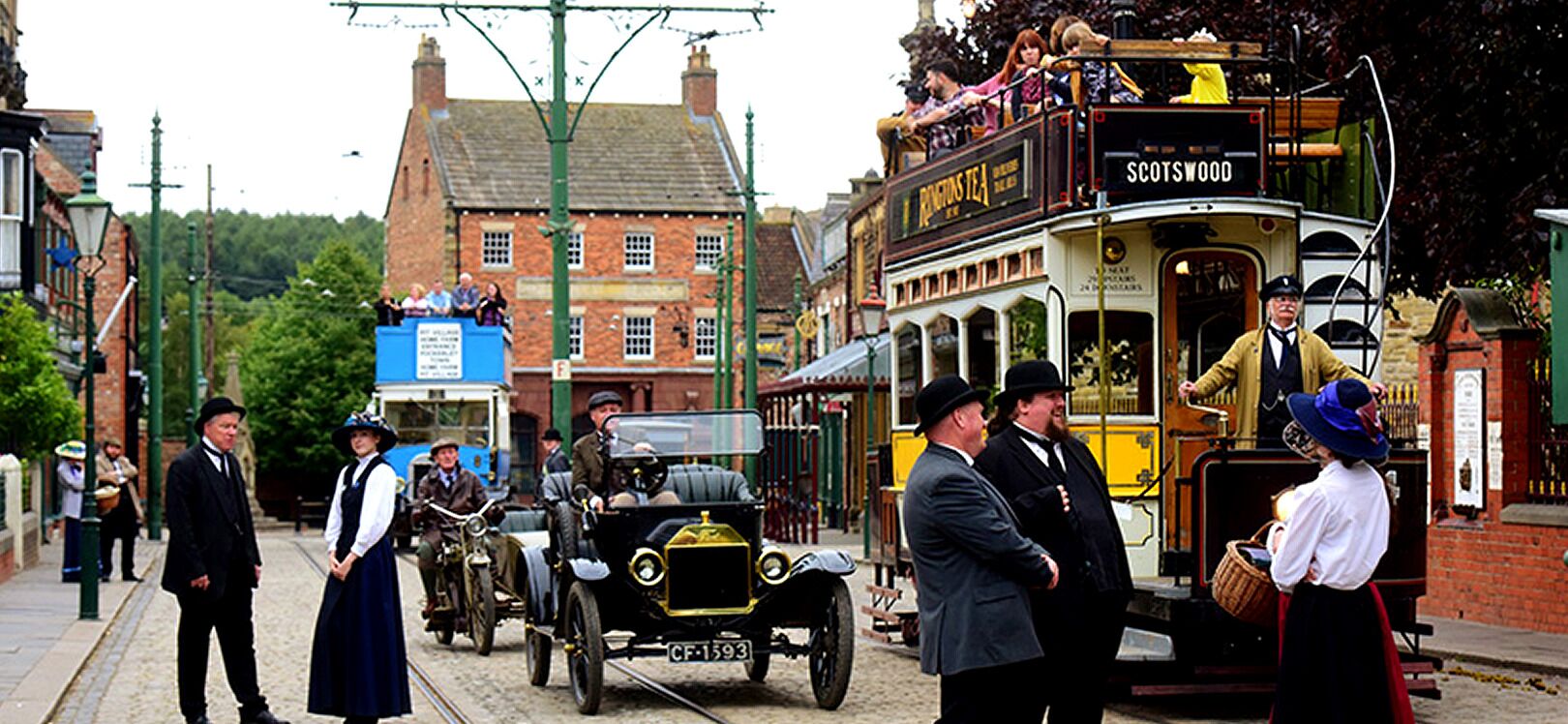 A vintage street scene with a double-decker bus, vintage cars, and people dressed in period clothing.