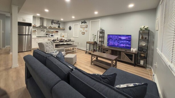 A modern living room with a grey sofa, a dining area with a kitchen in the background, and a TV displaying a cityscape. - Stylish Retreat with Fire Pit Near Casinos (Atlantic City)