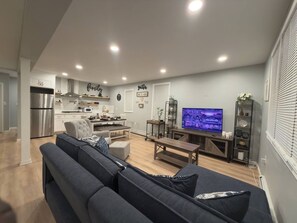 Een moderne woonkamer met een grijze bank, een eethoek en een keuken op de achtergrond, en een televisie waarop een stadsgezicht te zien is.
