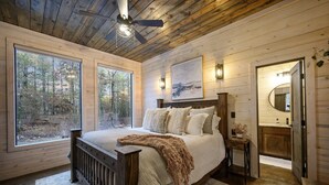Quarto com uma cama grande, teto de madeira e vista para a floresta por grandes janelas.