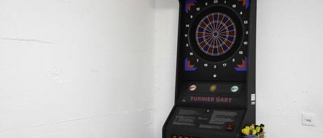 A dartboard mounted on a wall with a power outlet and a small basket of darts below it.