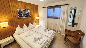 A hotel room with two beds, a wooden panel wall, a black and white photo, a window with curtains, a wooden chair, and a nightstand. - La Siala Apt.1 by Interhome (Falera)