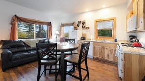 A cozy living room with a black leather sofa, wooden dining table, and chairs. A kitchen with wooden cabinets and a red stove is visible.