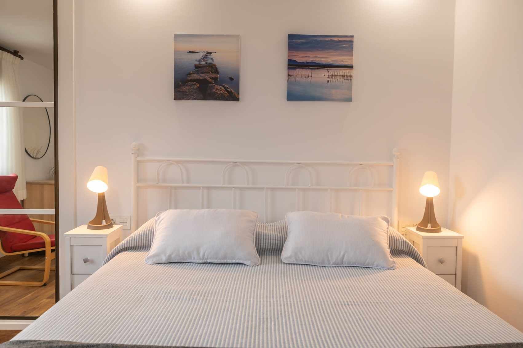 Un lit avec une literie blanche et deux oreillers, flanqué de deux tables de chevet avec des lampes, dans une pièce dotée d’une tête de lit blanche et de deux tableaux encadrés au mur.