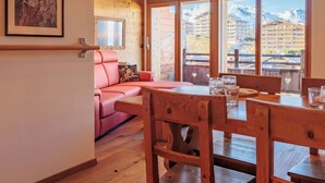 Une pièce chaleureuse avec un plancher en bois, un canapé rose, une table à manger en bois avec des chaises, et de grandes fenêtres offrant une vue sur une station de montagne.