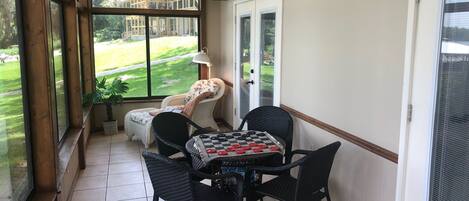 Una habitación soleada con vistas a un césped verde, un ventilador de techo, una mesa con mantel a cuadros y sillas de mimbre.