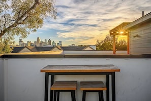 A rooftop terrace with a table and two chairs, overlooking a city skyline at sunset.