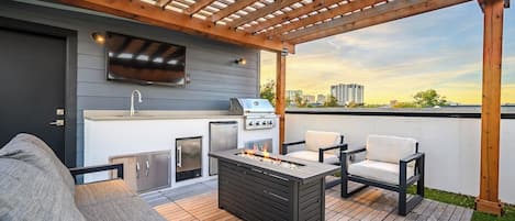 Un patio al aire libre con una parrilla, asientos, un televisor y vistas del horizonte de la ciudad.