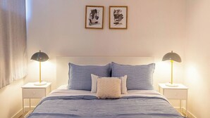Ein ordentlich gemachtes Bett mit blauer Bettdecke und Kissen, begleitet von zwei Nachttischen mit Lampen, und zwei gerahmten Kunstwerken an der Wand.