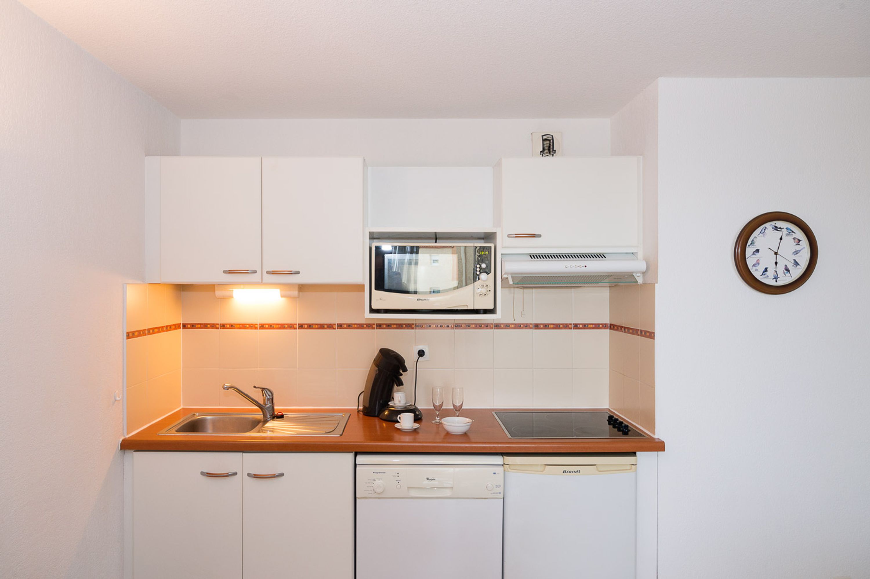 A compact kitchen with white cabinets, a microwave, a coffee maker, and a clock on the wall.