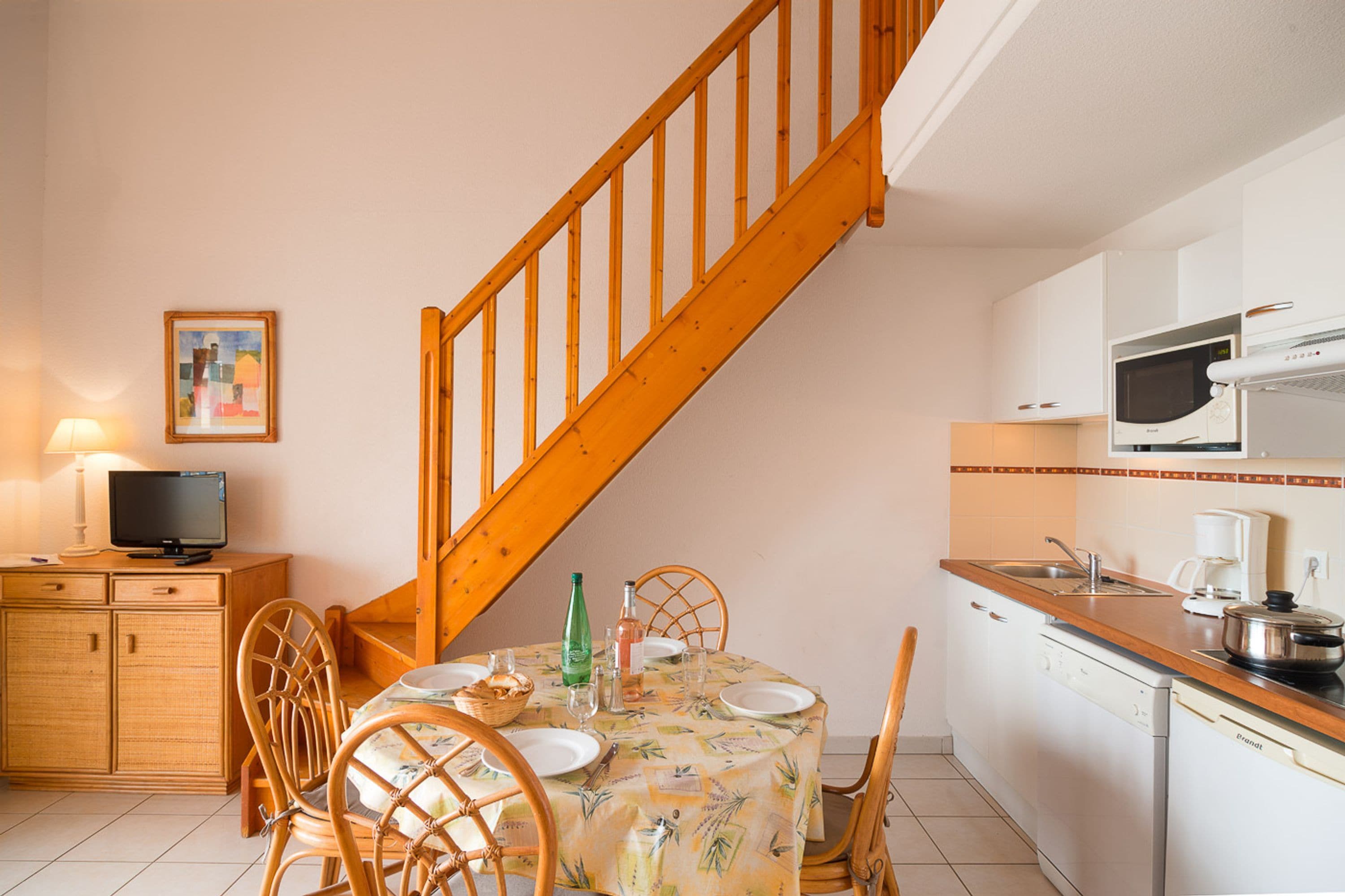 A compact kitchen with a dining area, wooden staircase, and a television on a wooden cabinet.