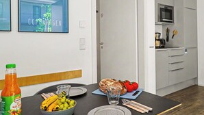 Een eethoek met een tafel voor twee personen, een fles sap en een brood. In de achtergrond is een keuken zichtbaar met moderne apparatuur.