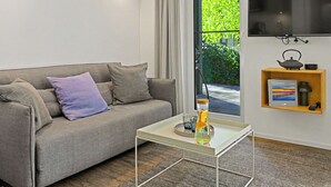 Een moderne woonkamer met een grijze bank, een glazen salontafel, een flatscreen-televisie aan de wand en een klein houten plankje met een boek en een waterkoker.