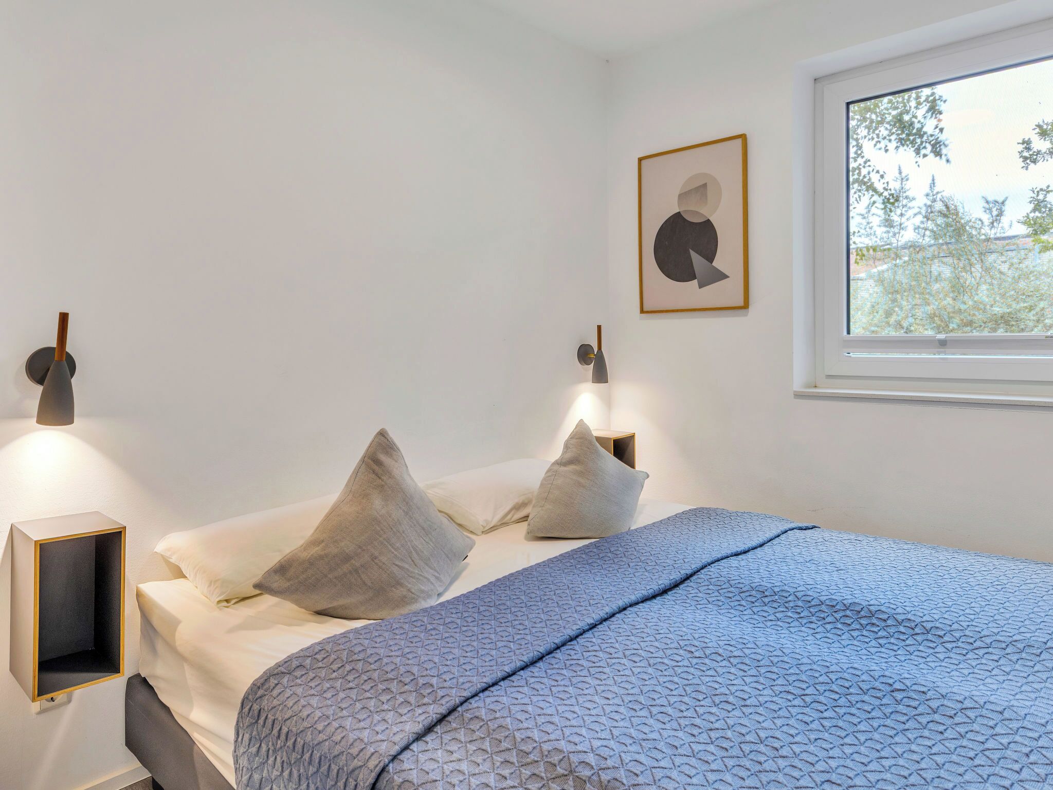 A bedroom with a bed, pillows, a bedside table, a wall-mounted lamp, and a window with a view of trees.