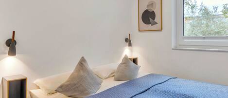 A bedroom with a bed, pillows, a bedside table, a wall-mounted lamp, and a window with a view of trees.
