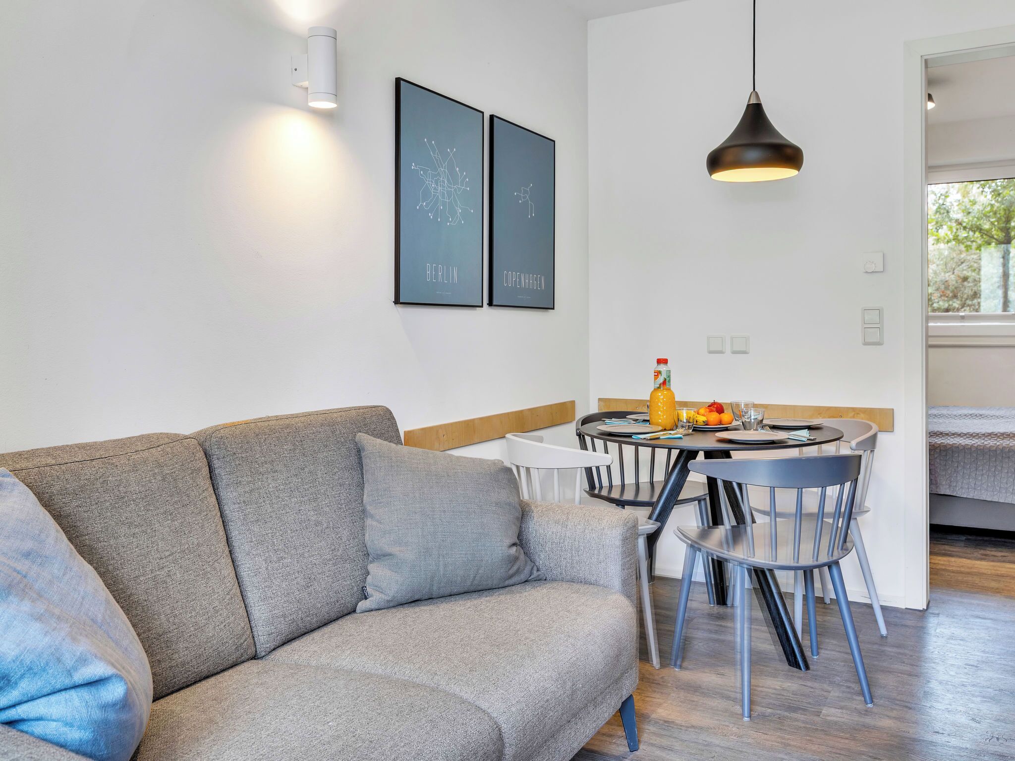 A modern living room with a grey sofa, a dining table with chairs, a hanging pendant light, and two framed pictures on the wall.