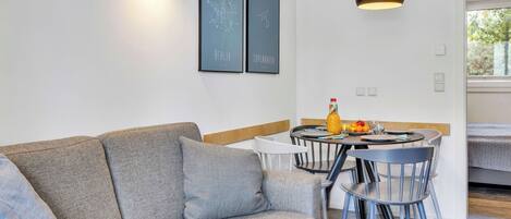 A modern living room with a grey sofa, a dining table with chairs, a hanging pendant light, and two framed pictures on the wall.