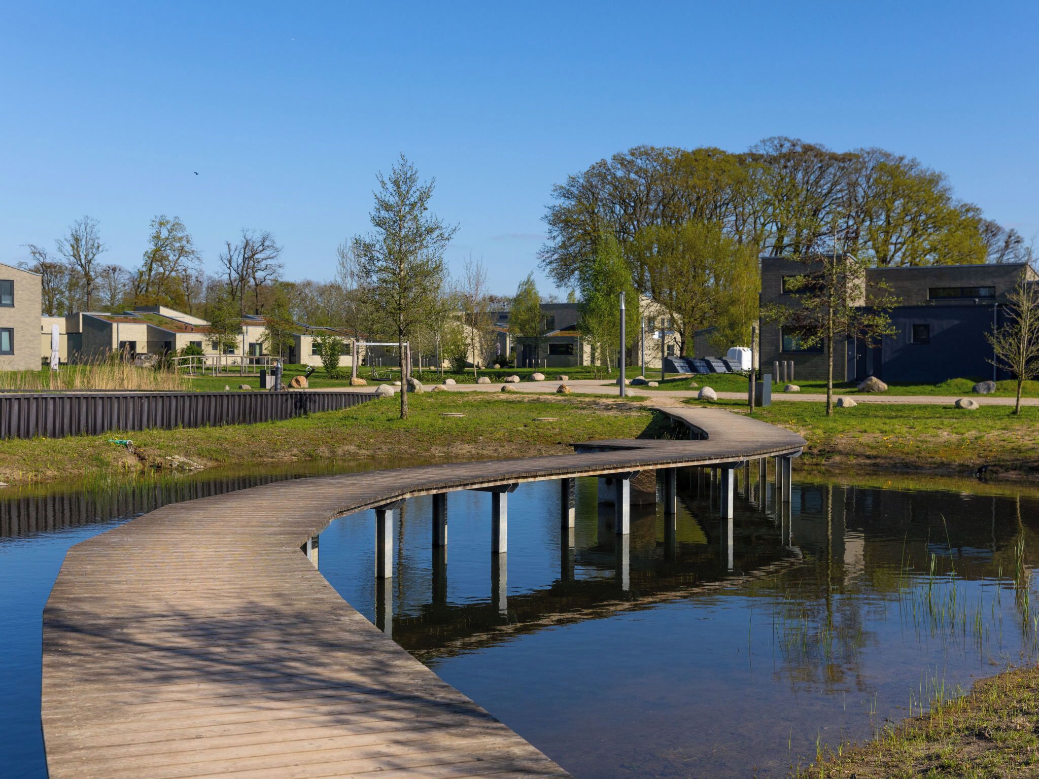 A wooden boardwalk over a calm water body, surrounded by greenery and modern buildings.