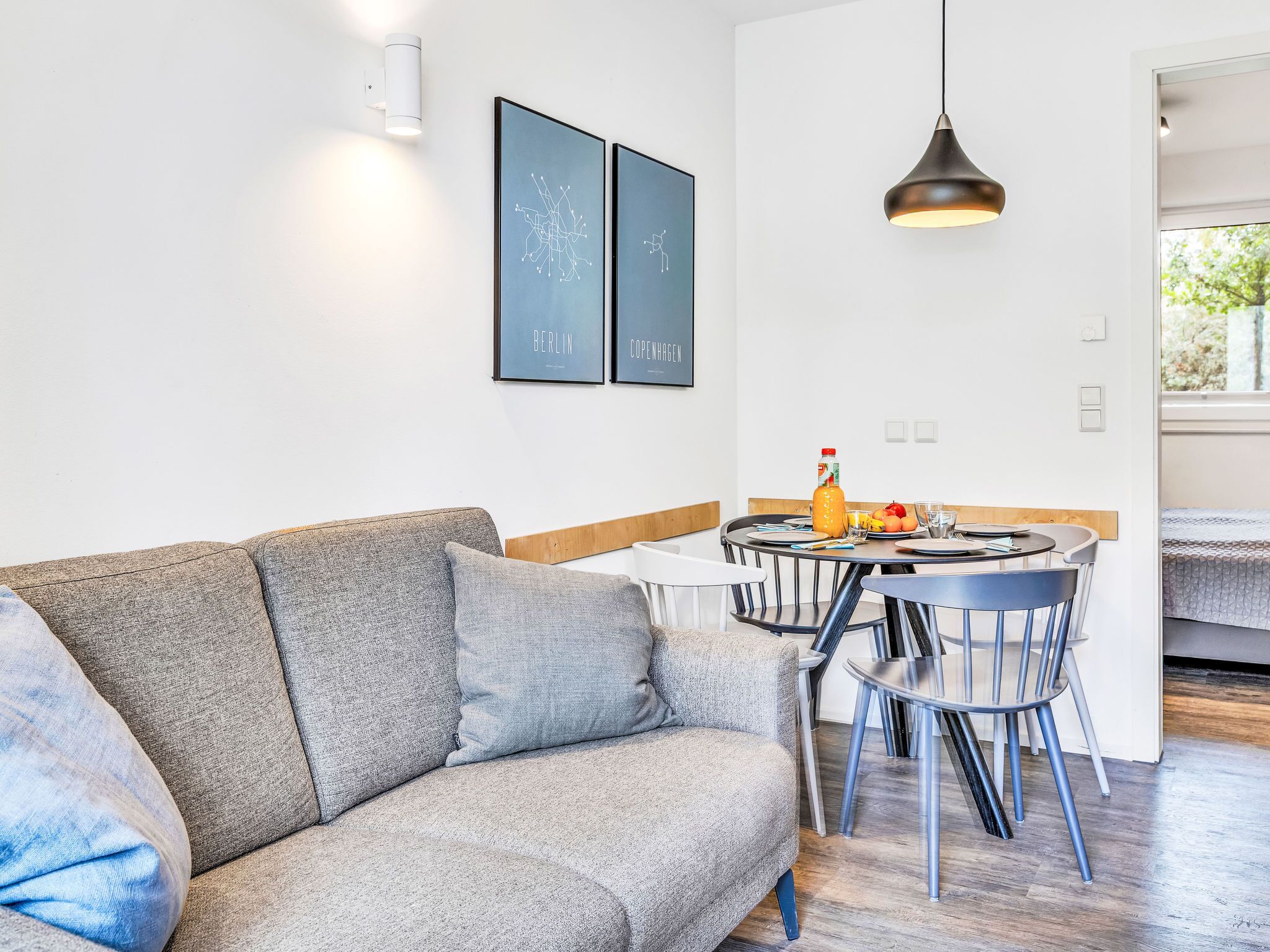 Een moderne woonkamer met een grijze bank, een ronde eettafel met stoelen, een hanglamp en twee ingelijste foto's aan de muur.