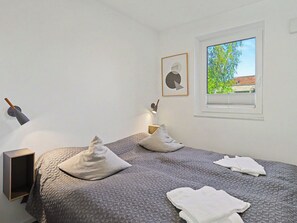 Ein Schlafzimmer mit Bett, Kissen, Nachttisch, Lampe, einem gerahmten Bild und einem Fenster mit Blick auf Grünflächen.