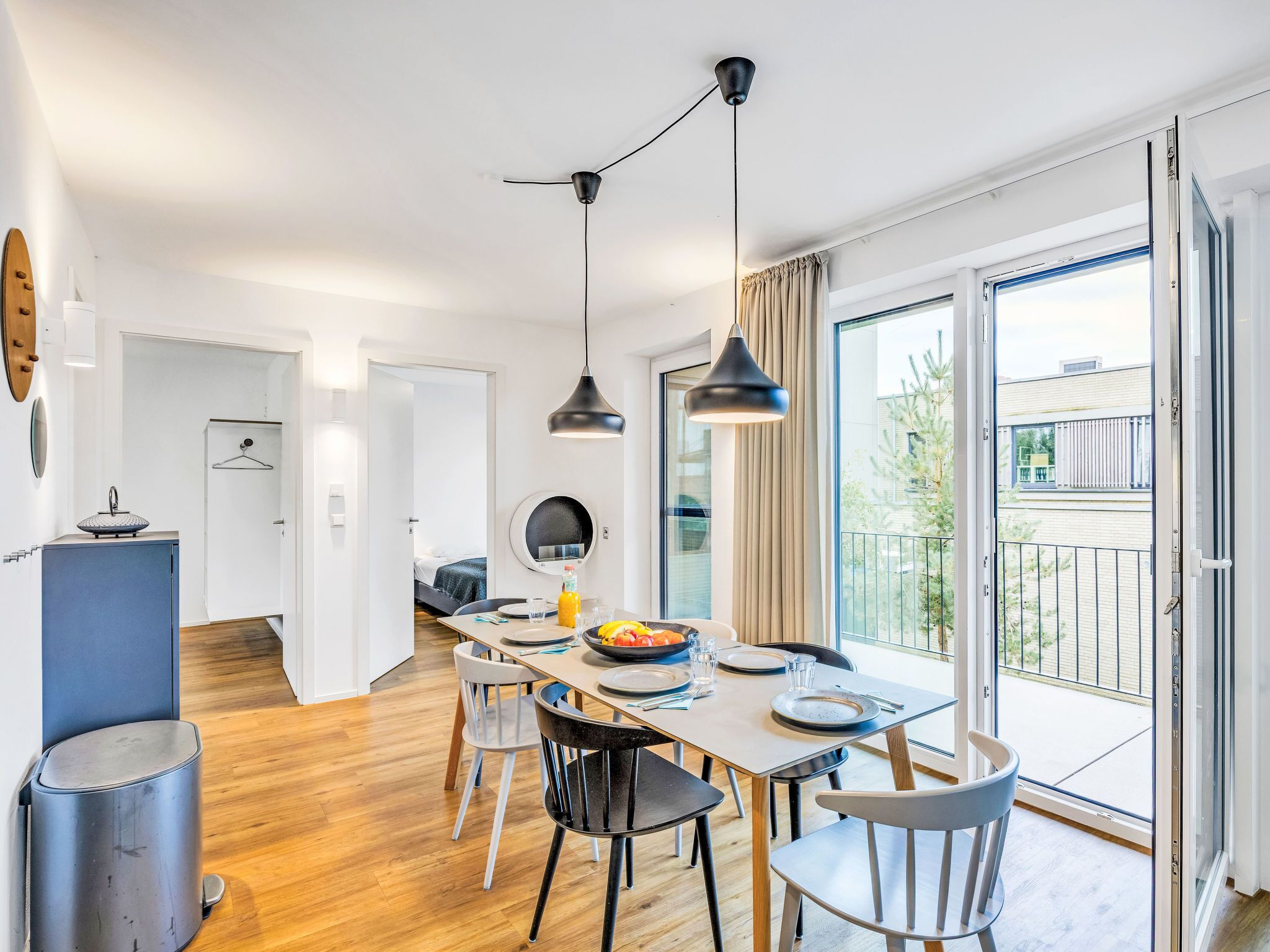 A modern dining area with a wooden table, chairs, and pendant lighting. A kitchen area is visible in the background.