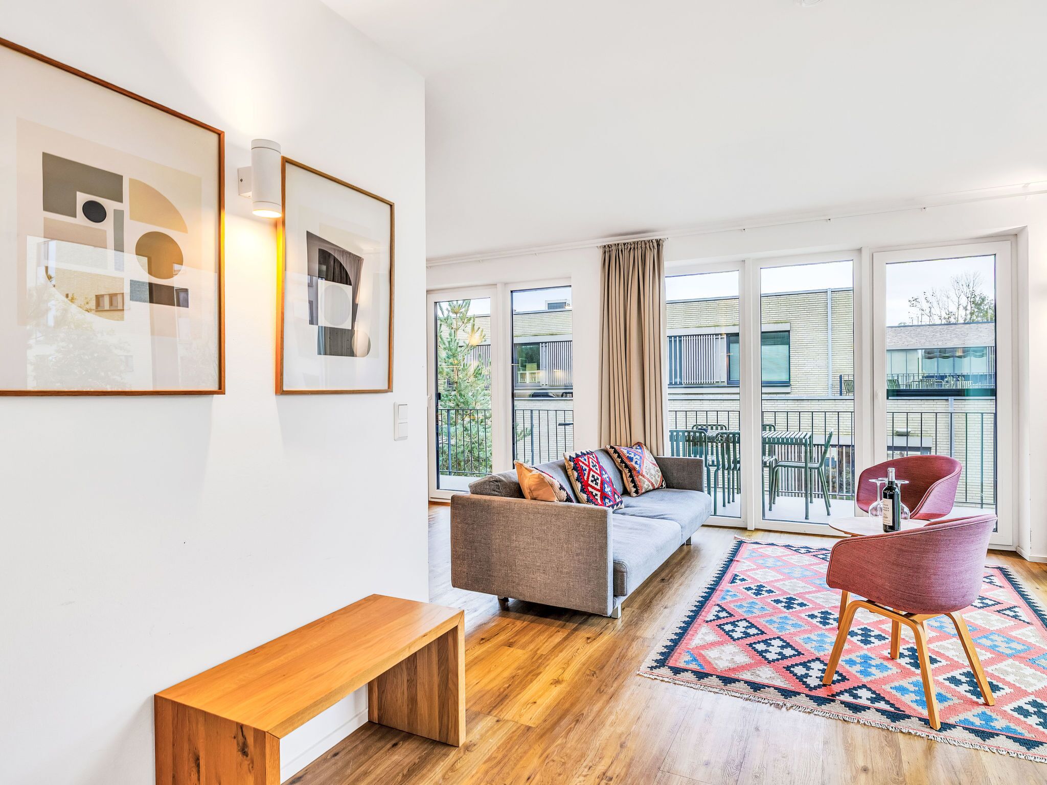 En moderne stue med sofa, bænk og en rød lænestol. Store vinduer og en balkon med havemøbler.