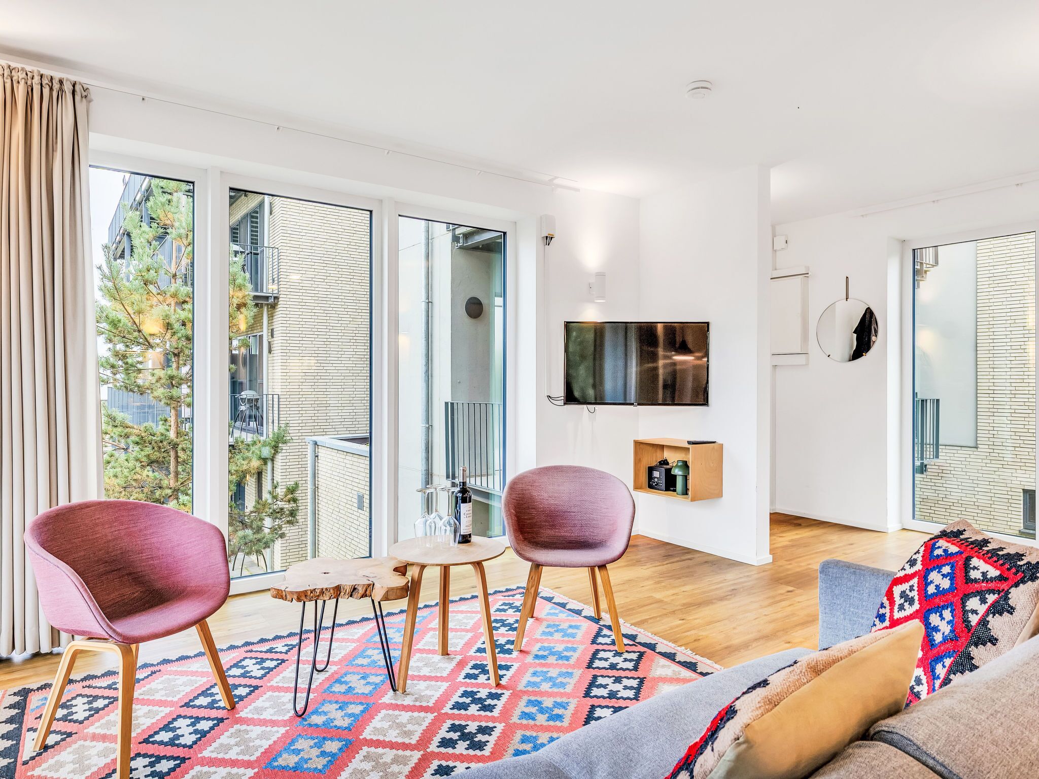 Una sala de estar moderna con ventanas amplias, un televisor de pantalla plana, un espejo redondo y una zona de estar acogedora con un tapete estampado.