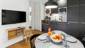 A modern kitchen with dark cabinets, a microwave, and a kettle. There is a dining table with plates, bowls, and a fruit bowl. A television is mounted on the wall, and a bookshelf is visible.