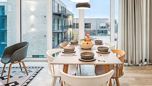 A modern dining area with a large table, multiple bowls, and a bottle of sauce. The room has large windows and a view of the city.