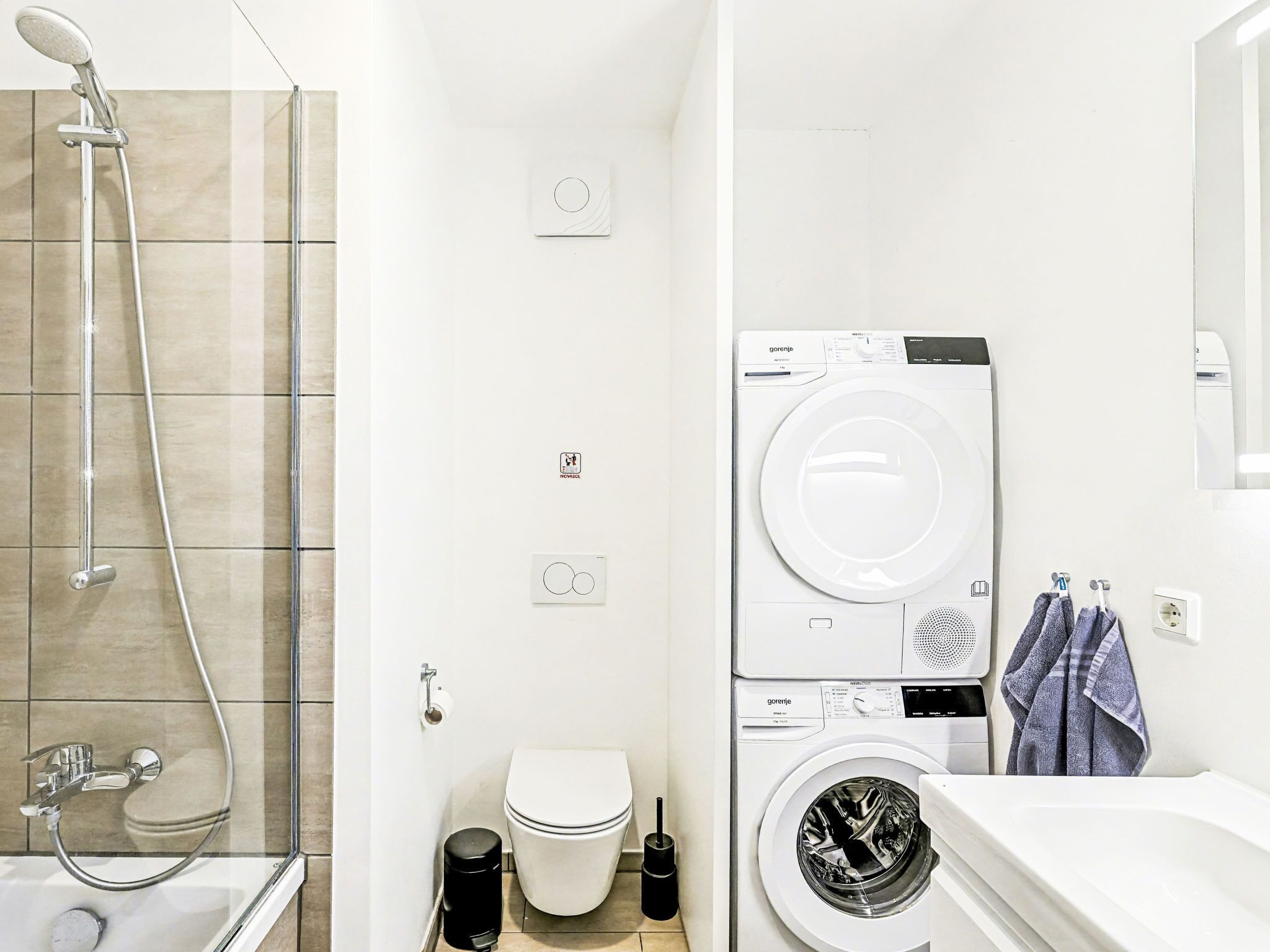 Een moderne badkamer met een douchecabine van glas, een toilet, een wasmachine en een wastafel met een handdoek aan de muur.