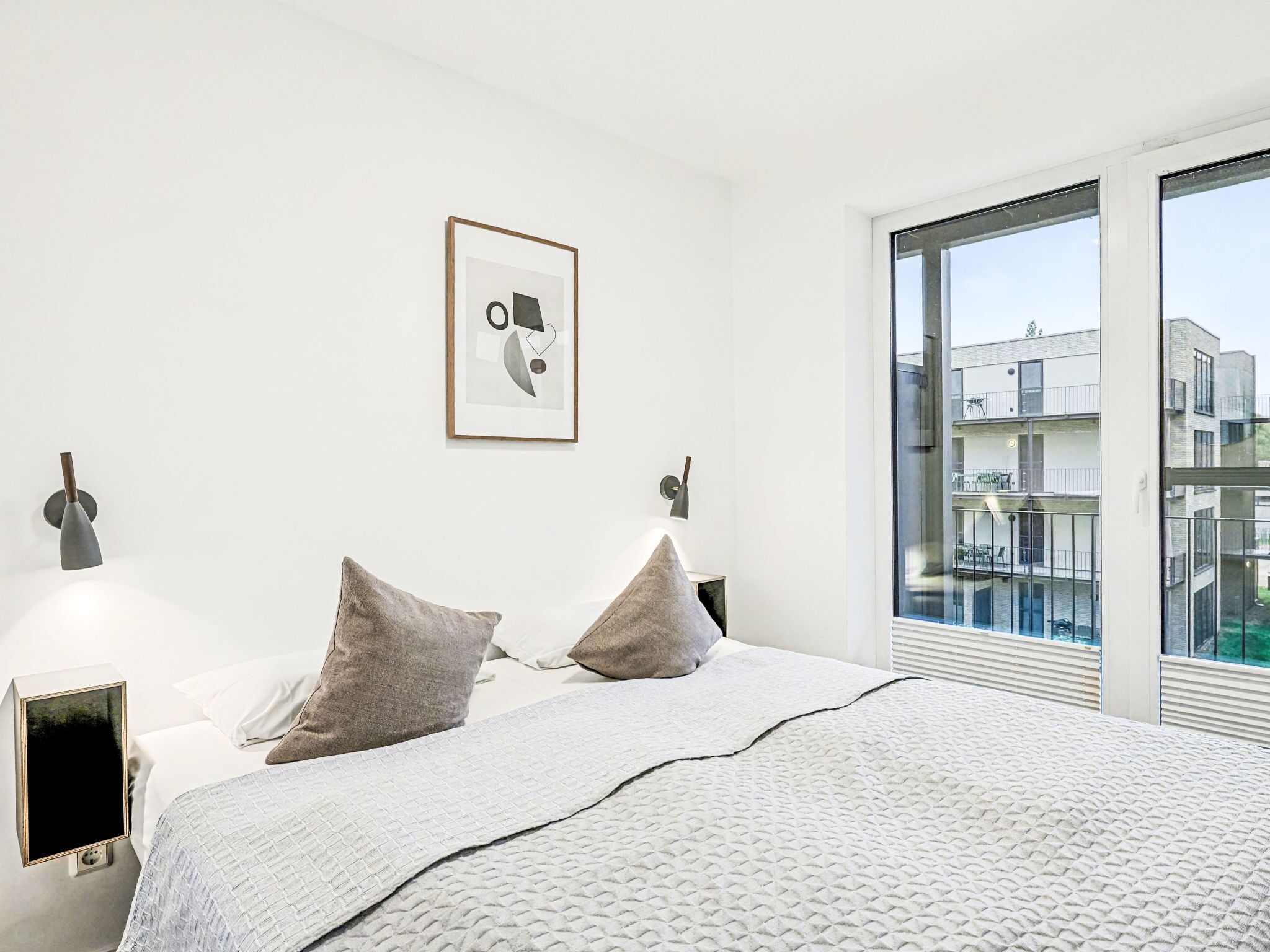 Ein modernes Schlafzimmer mit einem großen Bett, zwei Nachttischlampen, einem gerahmten Bild an der Wand und Blick auf Gebäude durch ein großes Fenster.