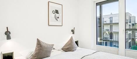 Ein modernes Schlafzimmer mit einem großen Bett, zwei Nachttischlampen, einem gerahmten Bild an der Wand und Blick auf Gebäude durch ein großes Fenster.