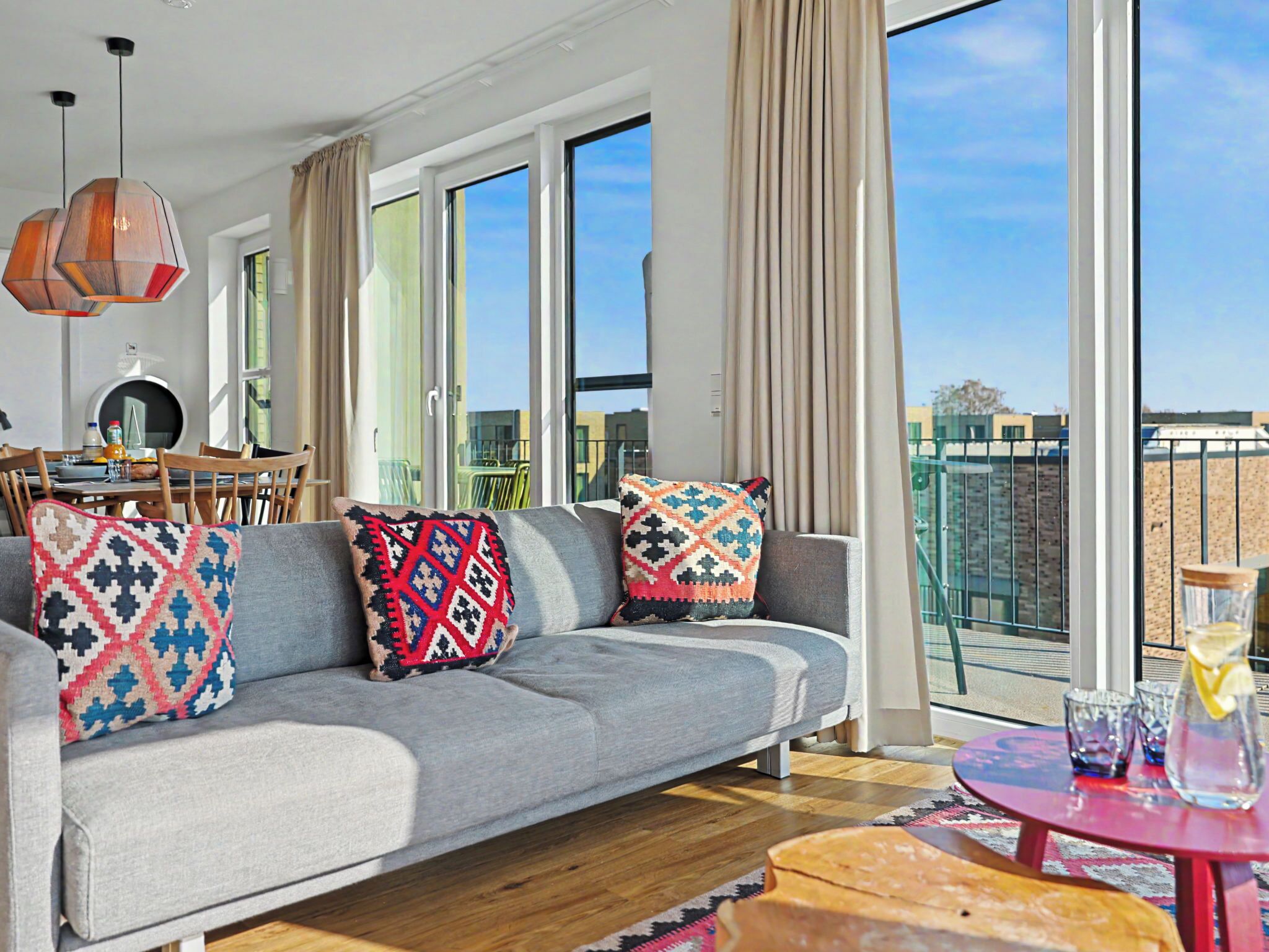 Un salon moderne avec un canapé gris, des coussins à motifs, une table à manger avec des chaises et de grandes fenêtres offrant une vue sur la ville.