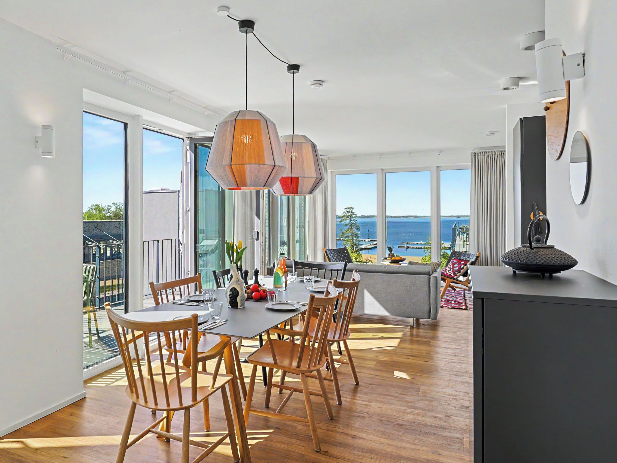 A modern dining area with a view of the sea, wooden chairs, a large table, and pendant lighting.