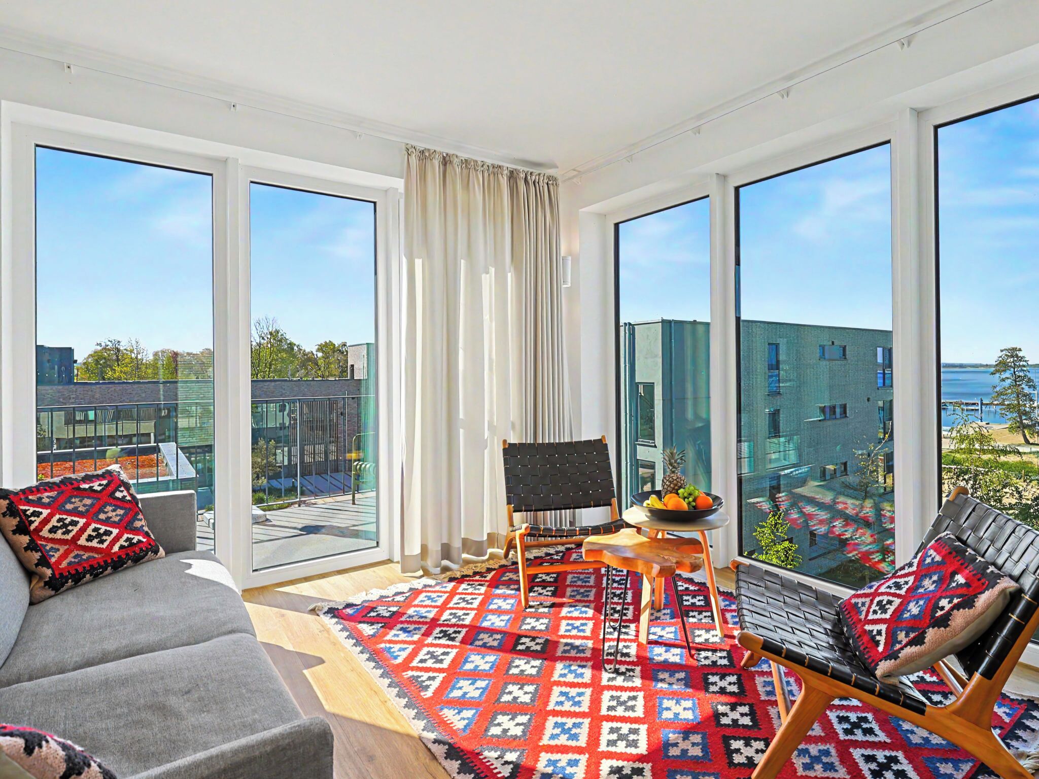 A modern living room with a sofa, a chair, a small table with fruit, and large windows offering a city view.