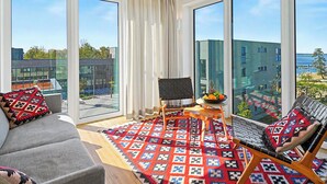 A modern living room with a sofa, a chair, a small table with fruit, and large windows offering a city view.