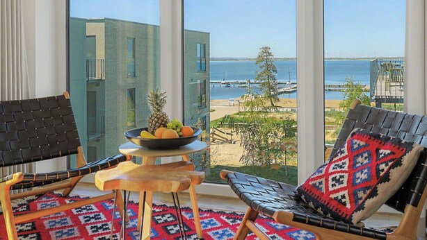 A balcony with two chairs, a small table with fruit, and a view of the sea and buildings.