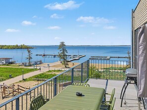 A balcony with a view of a lake, a grill, and a table with chairs.