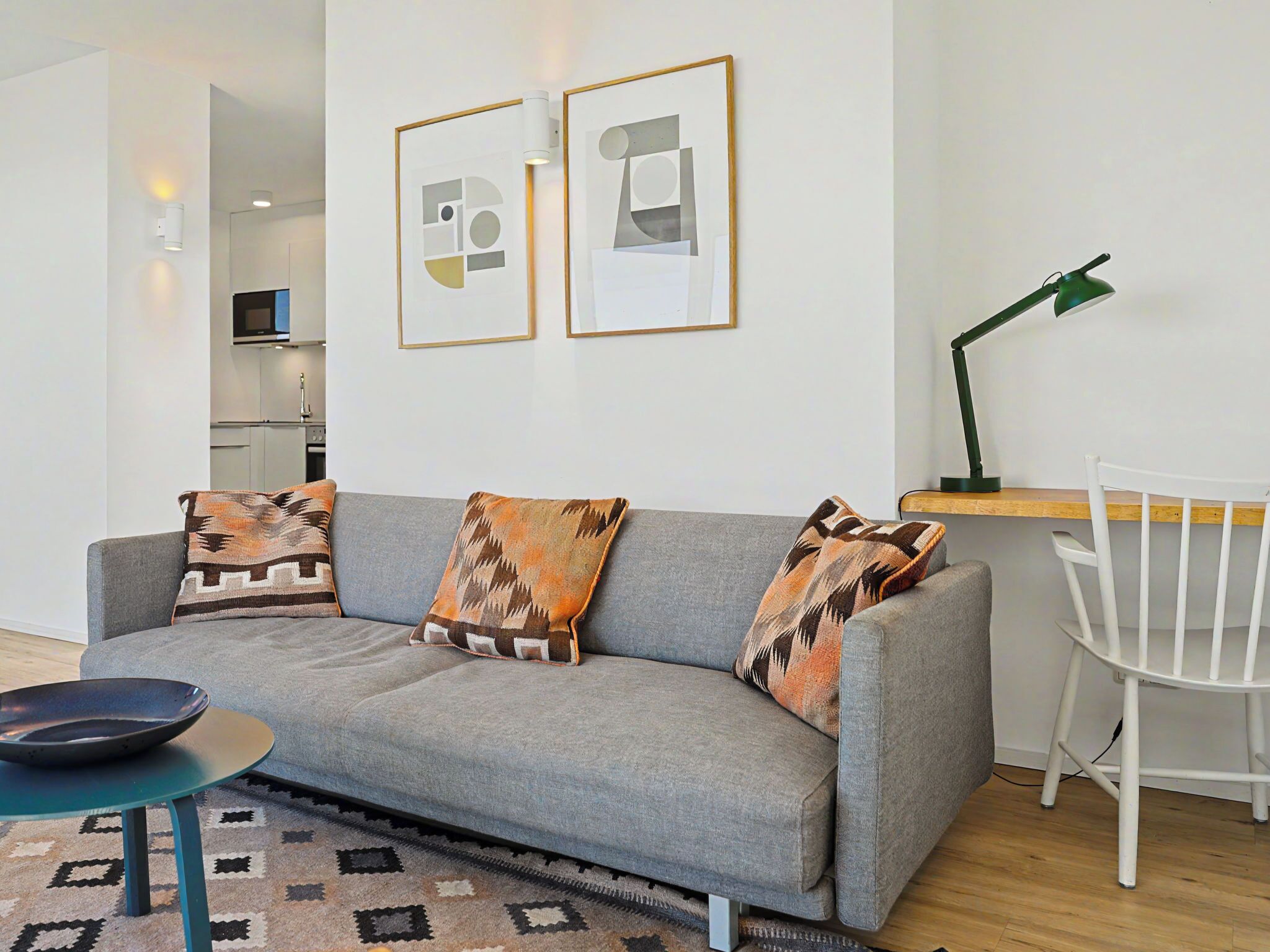 A living room with a grey sofa, patterned cushions, a small round table, a white chair, and two framed abstract artworks on the wall.
