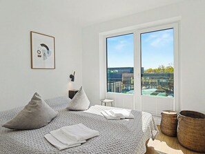 A modern bedroom with a large bed, two pillows, a bedside table with a lamp, a framed picture, and a view of a building complex through a sliding door.