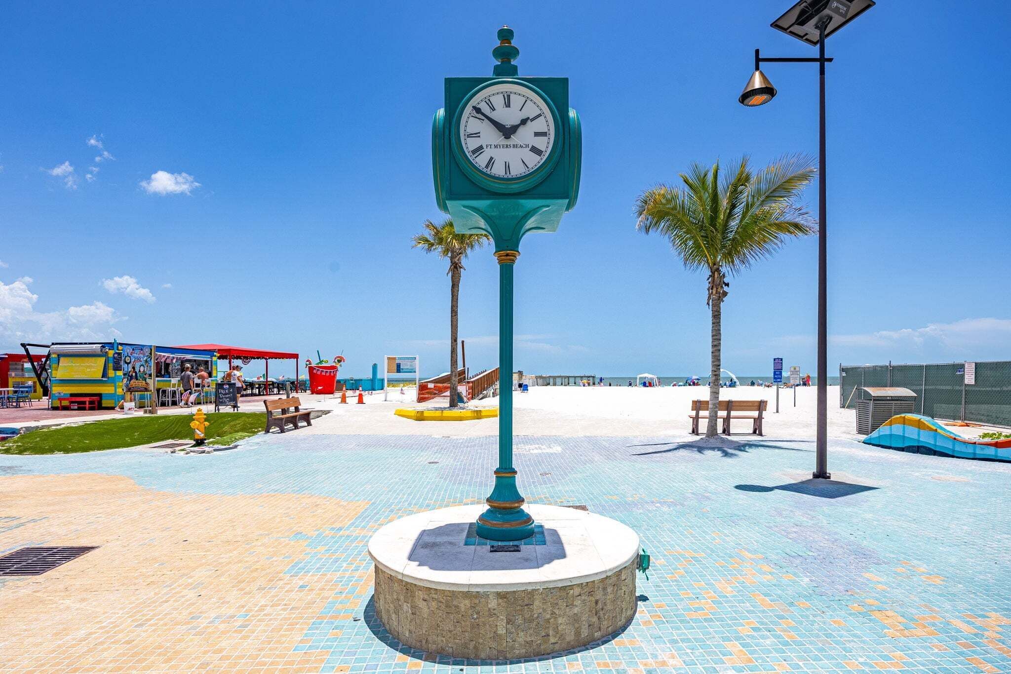 Um relógio com mostrador branco e números pretos, fixado num poste verde-água com base circular, situado numa passarela de azulejos com palmeiras e guarda-sóis de praia ao fundo.