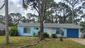 A single-story house with a blue exterior, a garage, and a front yard with grass and trees. - That cute house on Emerald! (Palm Bay)