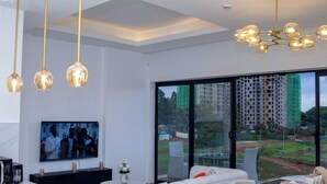A modern living room with a large TV, white sofas, a glass dining table, and a view of a cityscape through large windows. - 🌇 Fully Serviced 1-Bedroom Apartment at Riverbank, Two Rivers – Nairobi (Nairobi)