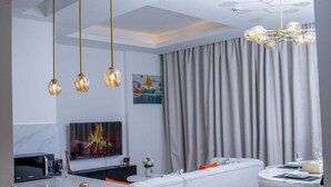A modern living room with a white sofa, a dining table with chairs, a kitchen area with a microwave, and a large window with curtains. - 🌇 Fully Serviced 1-Bedroom Apartment at Riverbank, Two Rivers – Nairobi (Nairobi)